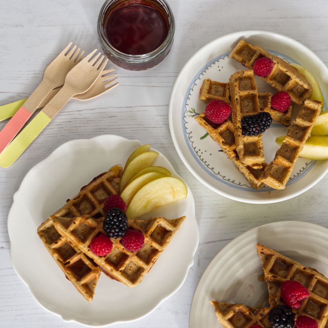 Golden buckwheat waffles with fresh raspberries, blackberries, and apple slices on white plates.