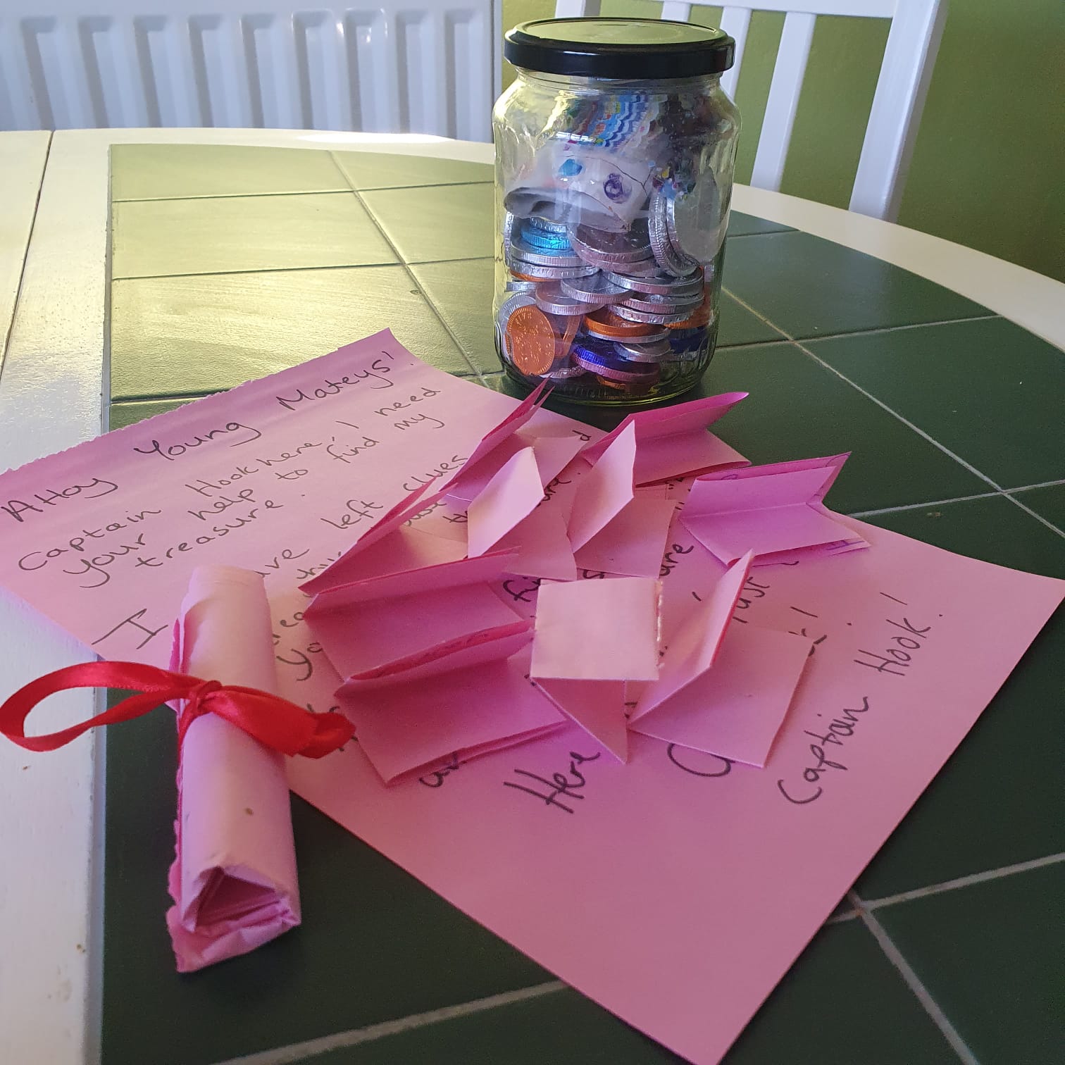 Pink treasure hunt clues and a jar of coins on a green and white table.