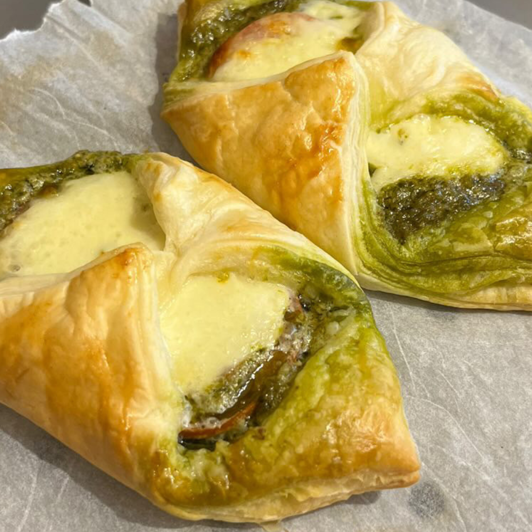 Two golden-brown pesto puff pastries with melted cheese and tomato on baking paper.