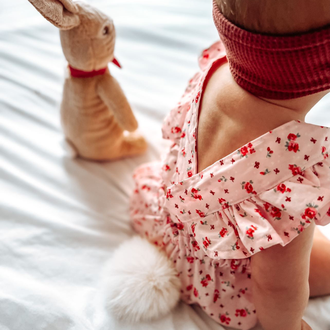 Baby in a pink floral romper with a fluffy white bunny tail, next to a toy bunny.