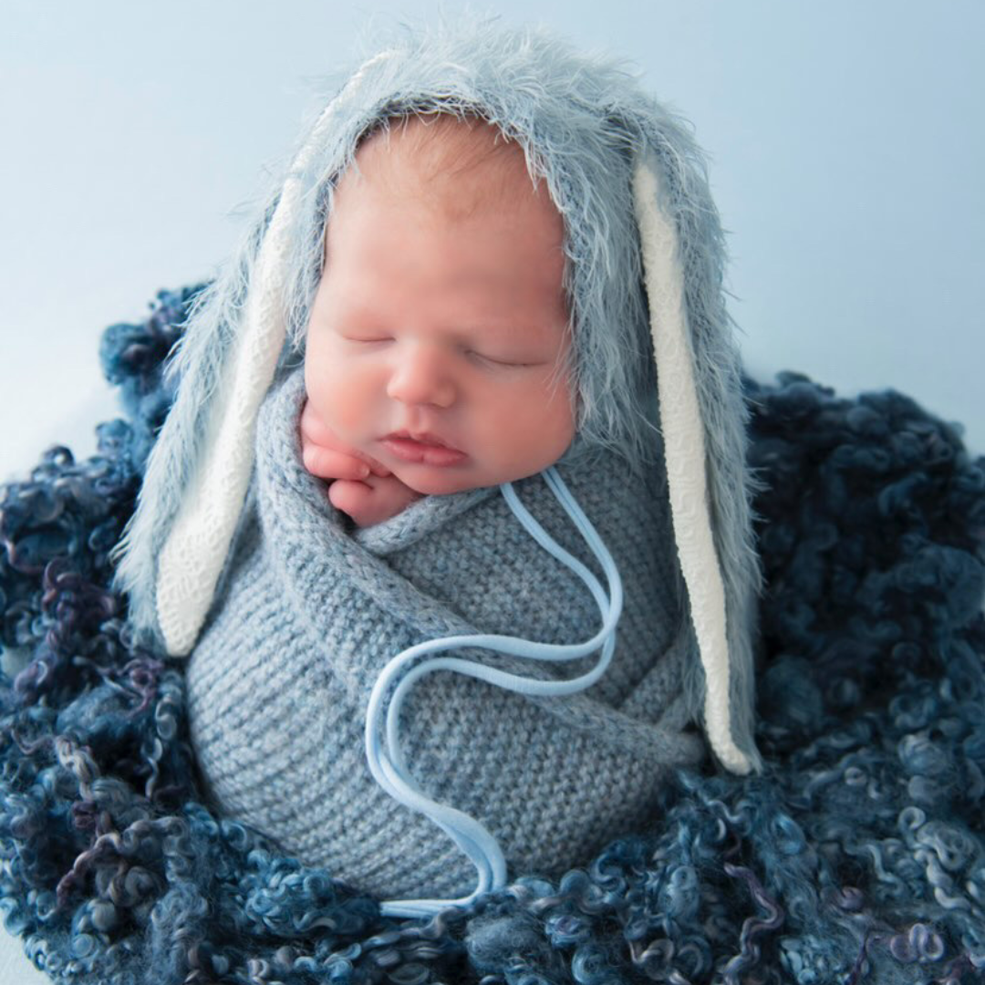 Newborn baby asleep in a blue knitted wrap, wearing a soft blue bunny bonnet with white ears.