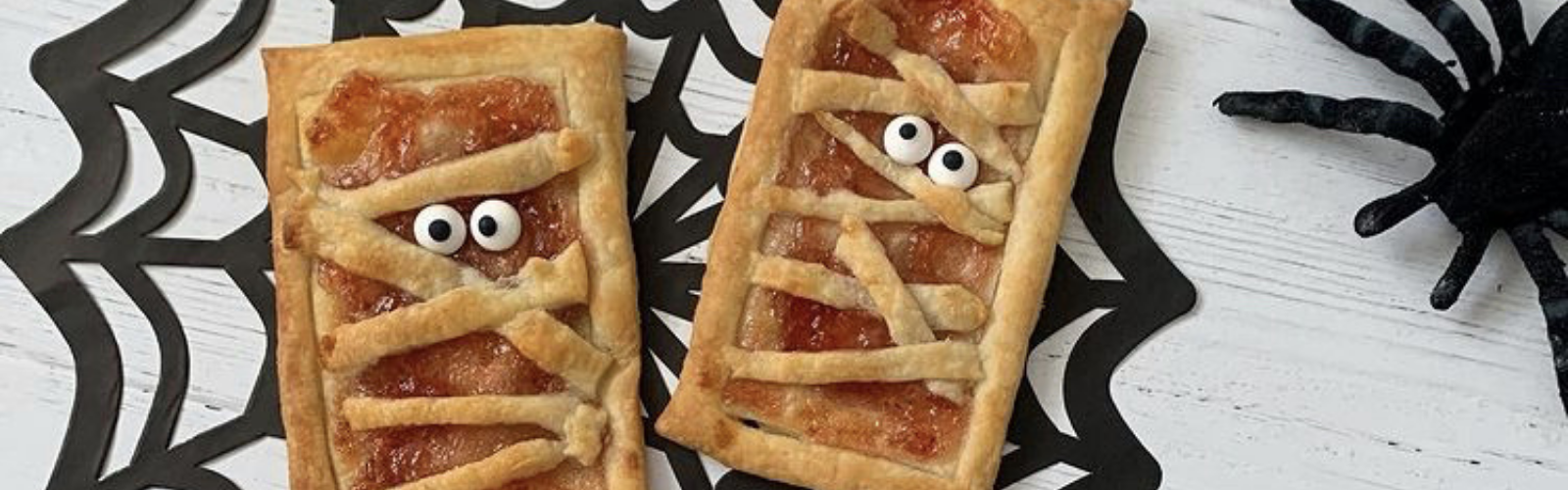 Two golden-brown jammy mummy pastries with googly eyes on a black spiderweb mat.