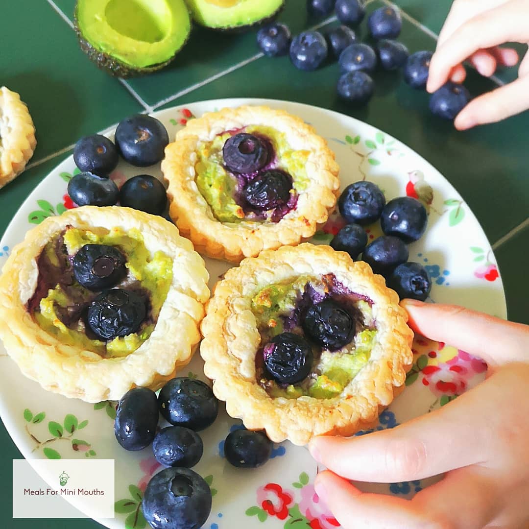 Three golden puff pastry tarts with green filling and fresh blueberries on a floral plate.