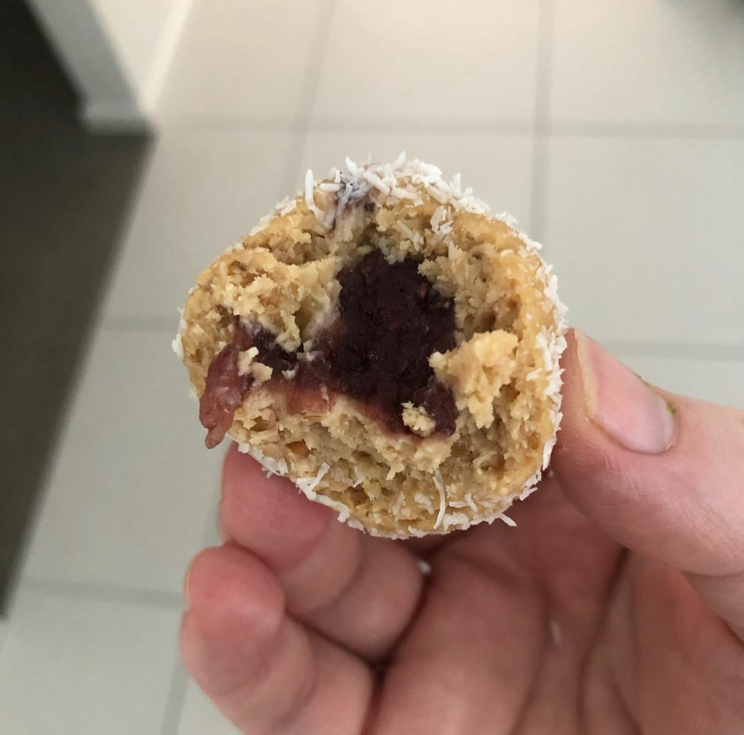 Hand holding a peanut butter bliss ball, cut open to reveal a dark chia jam centre and coconut coating.