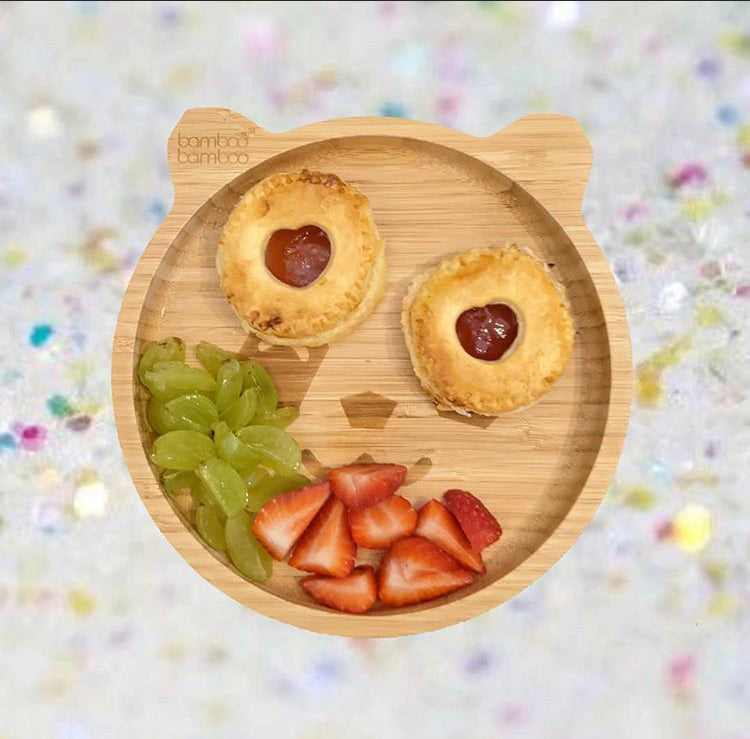 Two golden chia jam pastries with heart cut-outs, green grapes, and sliced strawberries on a bamboo plate.