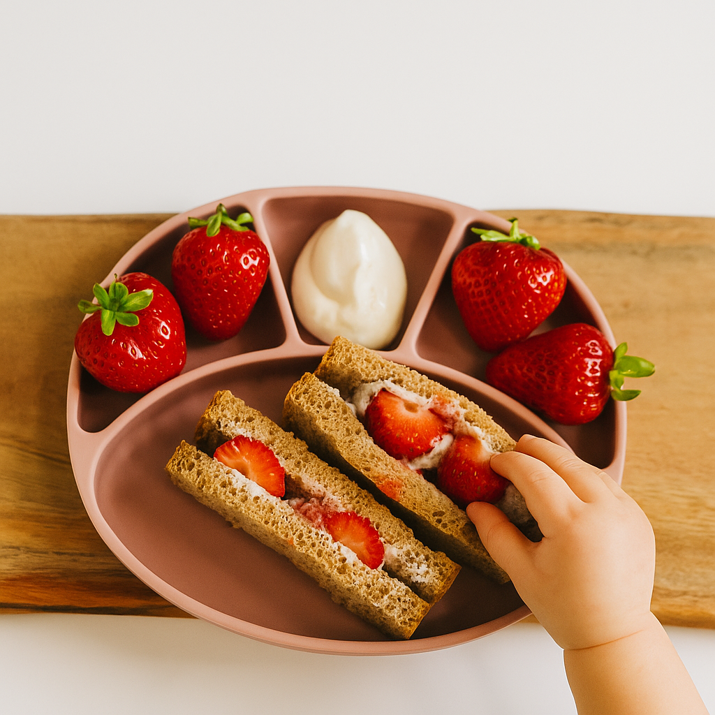 Weaning-friendly strawberry and cream sandwiches on a pink divided plate, with a baby's hand reaching for one.