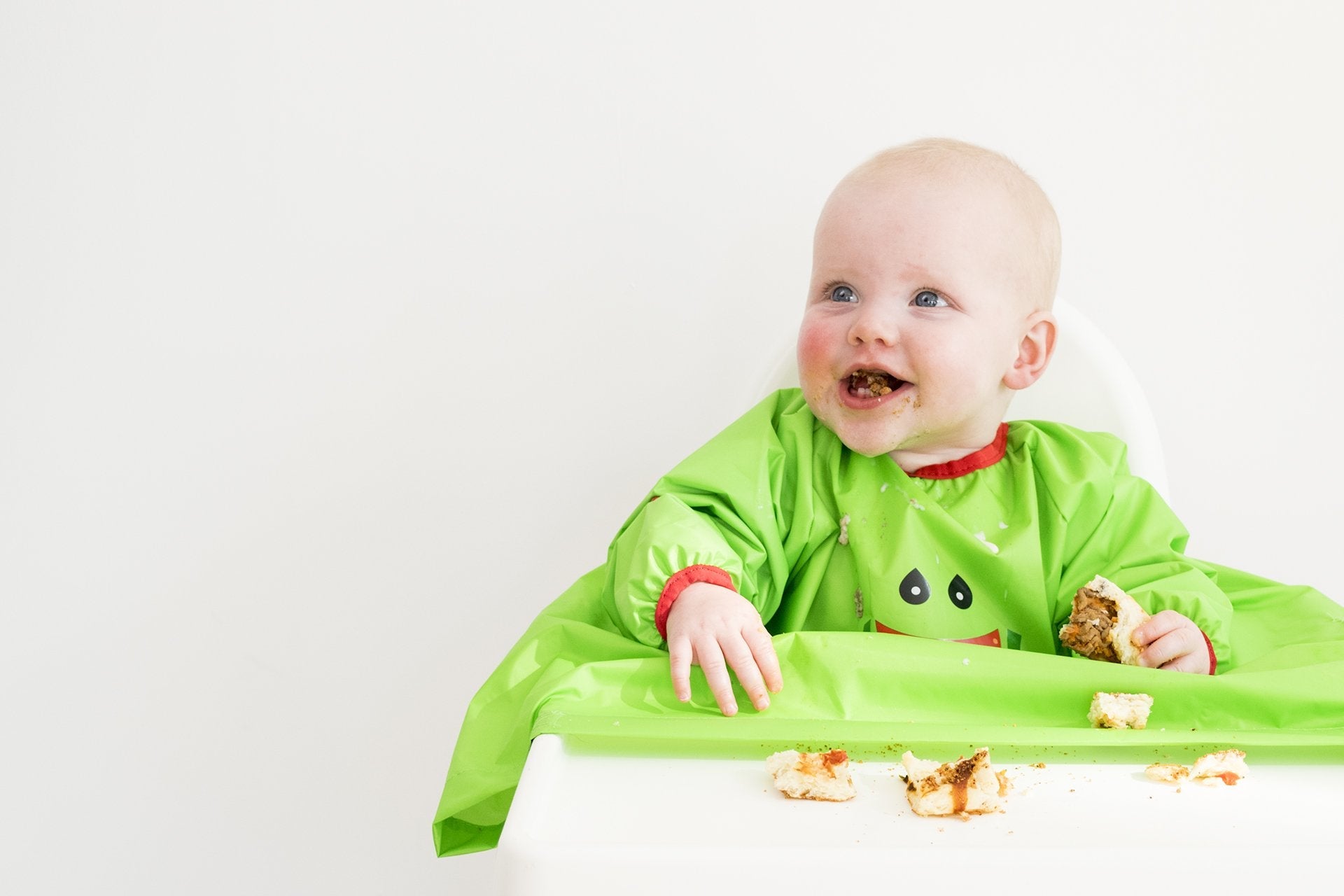 Happy baby in a green bib, messy from self-feeding pizza in a highchair.