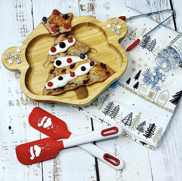 Christmas tree pancakes on a bamboo plate with festive sprinkles and spatulas.