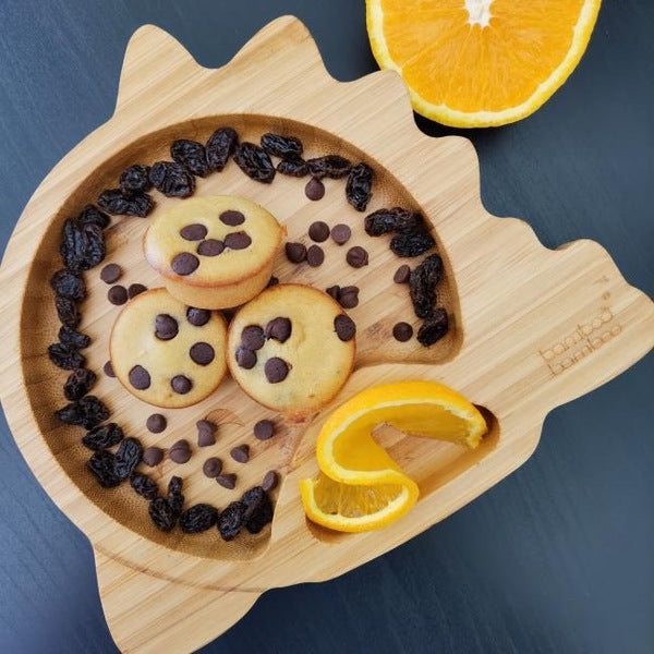Three mini muffins with chocolate chips on a bamboo plate, surrounded by raisins and orange slices.