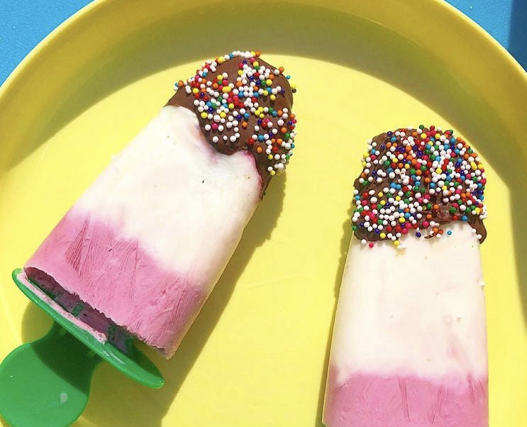 Two homemade pink and white yoghurt lollies with chocolate and colourful sprinkles on a yellow plate.