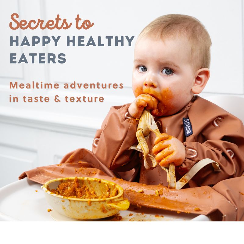 Messy baby in a brown bib exploring food with a fork and spoon, a yellow bowl of food nearby.