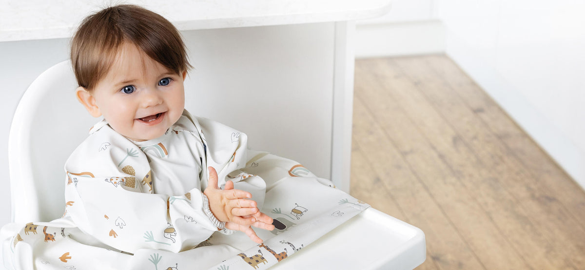 Happy baby in a long-sleeved, neutral-toned weaning bib with playful animal and rainbow patterns.