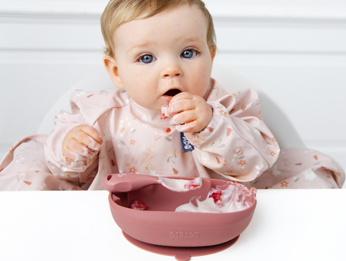 A baby wearing a light pink flower-patterned bib sits at a white high chair, reaching for food in a dusty rose silicone suction bowl.  - image