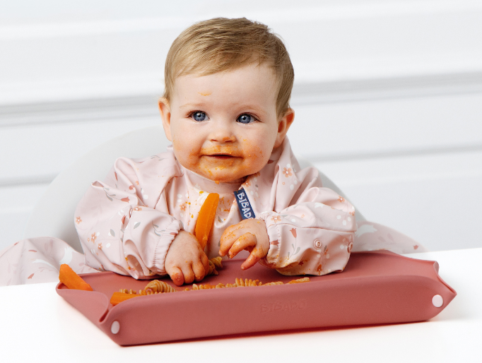A baby sitting in a high chair wearing a light pink bib with floral patterns, holding an orange carrot. The baby is resting on a dusty rose silicone bib tray on a white table against a white paneled wall. - image