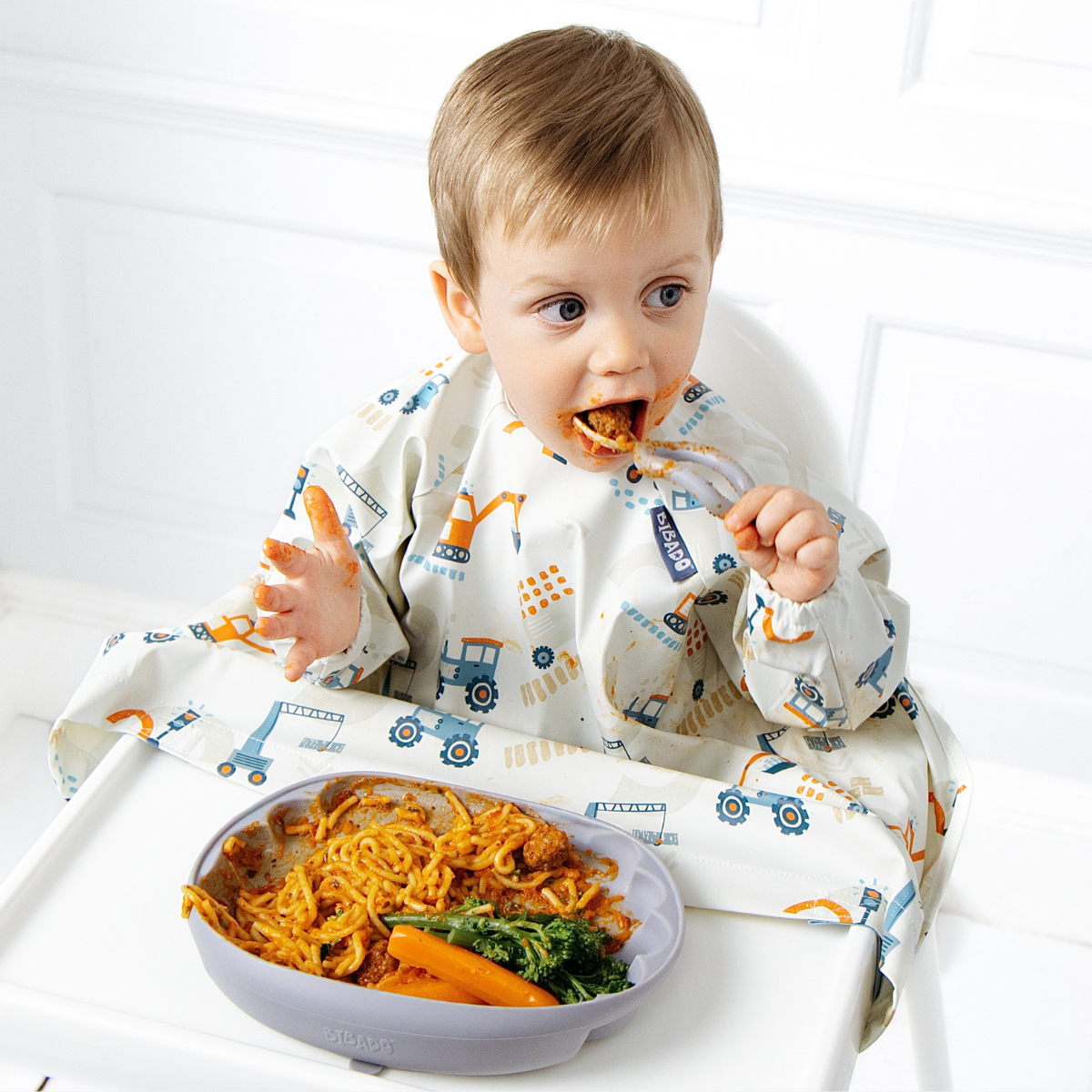 A baby wearing a white bib with blue and orange construction vehicle prints sits in a high chair, eating spaghetti with broccoli and carrots from a blue bowl on a white high chair. - Product info image
