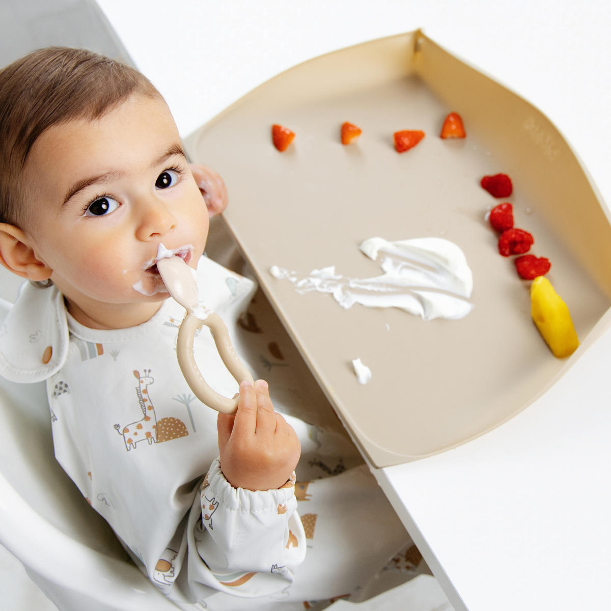 A baby wearing a white long-sleeve bib with a safari print sits in a high chair, eating yoghurt from a beige spoon. A matching beige feeding mat sits on the table, holding some fruit pieces and a smear of white yoghurt. - Product info image