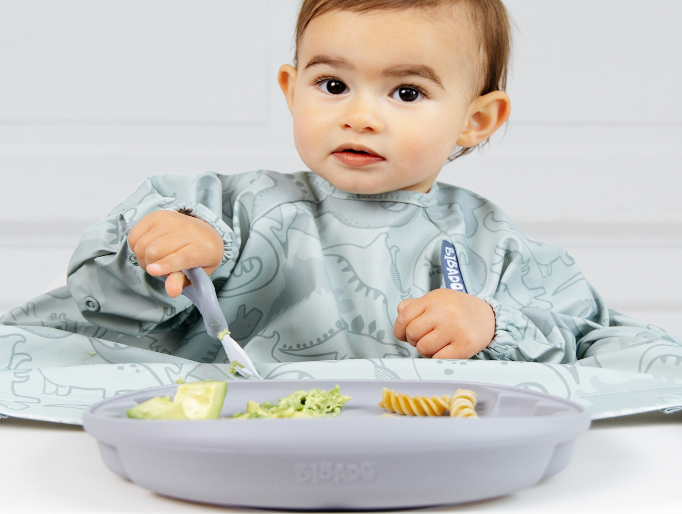 A baby wearing a light blue dinosaur-print bib sits at a table with a blue-grey silicone plate and a matching spork, eating pasta and avocado. - image