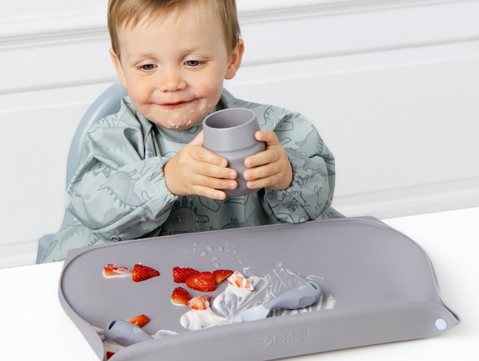 A toddler wearing a light blue dino patterned bib sits in a high chair, holding a blue cup. In front of them is a blue silicone feeding mst with spilled strawberries and yoghurt on a white table against a white panelled wall. - image