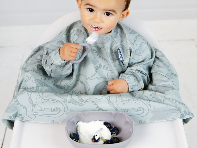 A baby sitting in a white high chair wearing a long-sleeve bib with a light blue-gray dinosaur pattern, eating yoghurt from a blue silicone bowl with matching Dippit spoon. - image