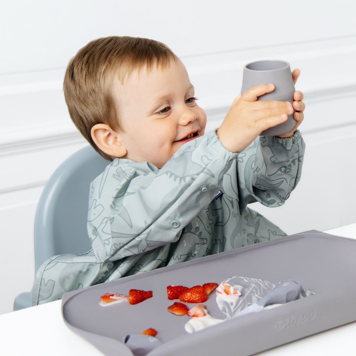 A smiling toddler in a light blue dinosaur-print bib sits in a high chair holding a blue-grey cup aloft. A matching feeding mat with food remnants is in the foreground on a white table against a white panelled wall. - Product info image