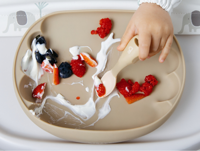 A close-up of a child's hand using a beige silicone baby spoon on a beige silicone scalloped plate, which is covered with yogurt, berries, and strawberries. The child is wearing a white long-sleeve bib with animal print. - image