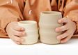Two beige silicone cups, one smaller than the other, held by a child wearing a brown Coverall bib. The child's hands and torso are visible, and the cups are shown against a clean white background.