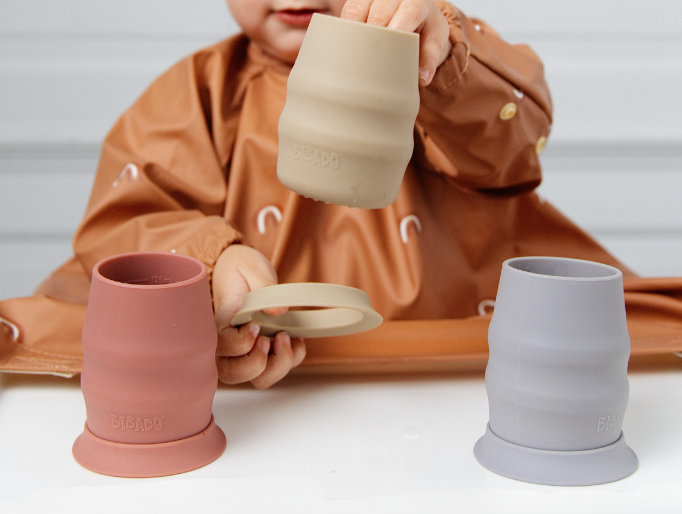 A toddler wearing a brown bib holds a beige silicone cup above two more cups on a white table: one dusty pink and one light blue.  - image