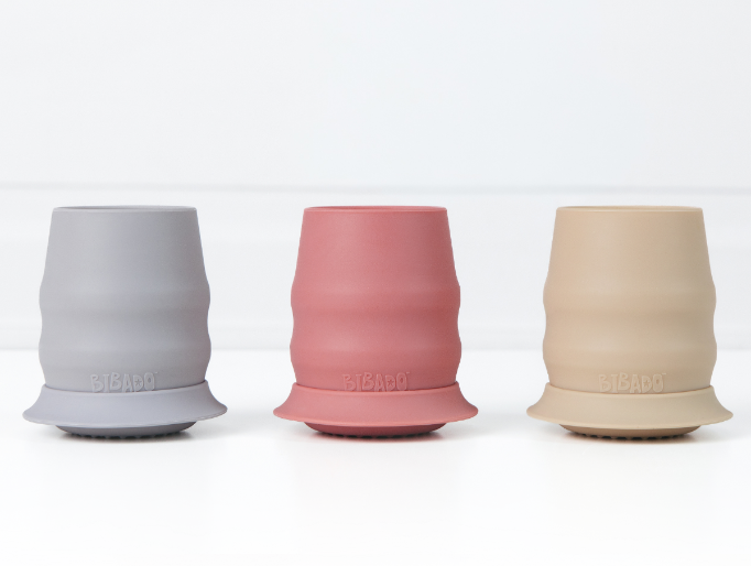 Three silicone baby cups with stabilising skirts in blue-grey, dusty pink, and beige, arranged side by side on a clean white surface. - image