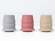 Three silicone baby cups with stabilising skirts in blue-grey, dusty pink, and beige, arranged side by side on a clean white surface.