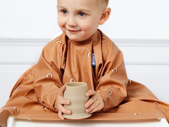 A toddler wearing a rust-brown bib and holding a beige cup, seated against a clean white background. - image