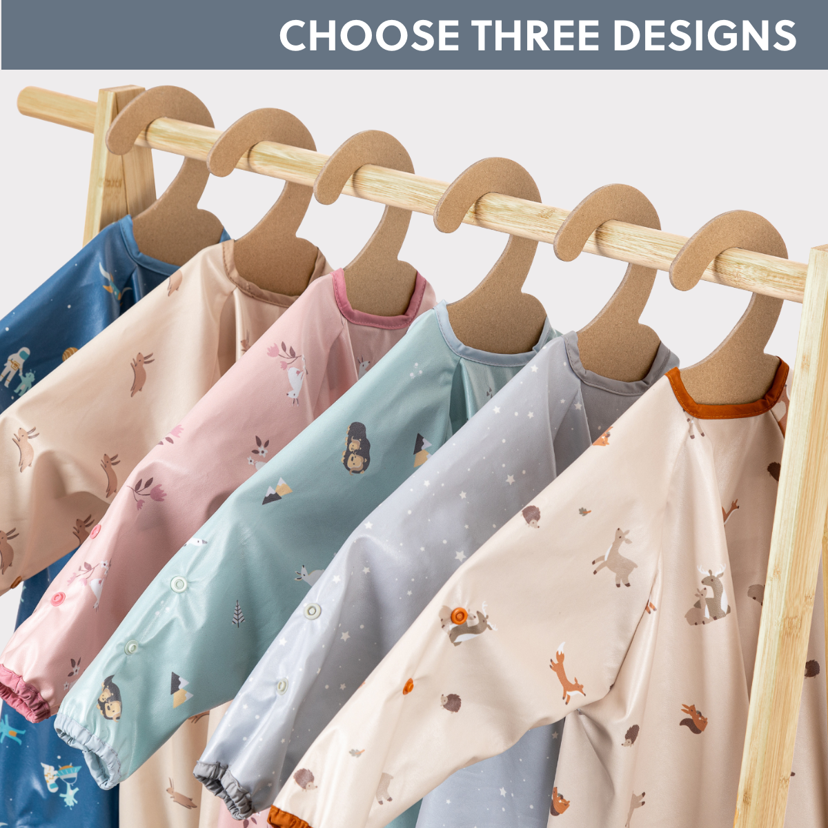 Six long-sleeve waterproof coverall weaning bibs in various heritage prints, hanging on a wooden rack. - Product image