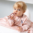 Blonde baby in a pink bib holding a rose-colored Dippit™ multi-stage baby spoon and dipper.