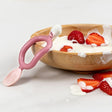 Pink silicone Dippit™ baby spoon and textured dipper in a wooden bowl with yogurt and strawberries.