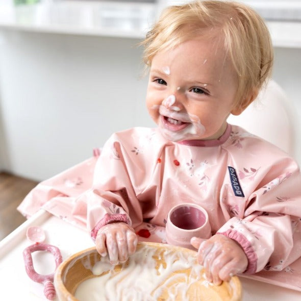 Happy baby in a pink floral bib, covered in yogurt, holding a wooden bowl and pink Dippit™ spoon. - Product image