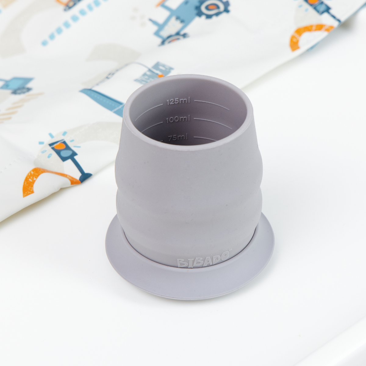 A gray silicone baby feeding cup with measurement markings inside, shown on a white surface with a patterned bib in the background. - Product image