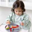 Child in a green Dino Coverall bib eating from a mist silicone divider plate with fruits.