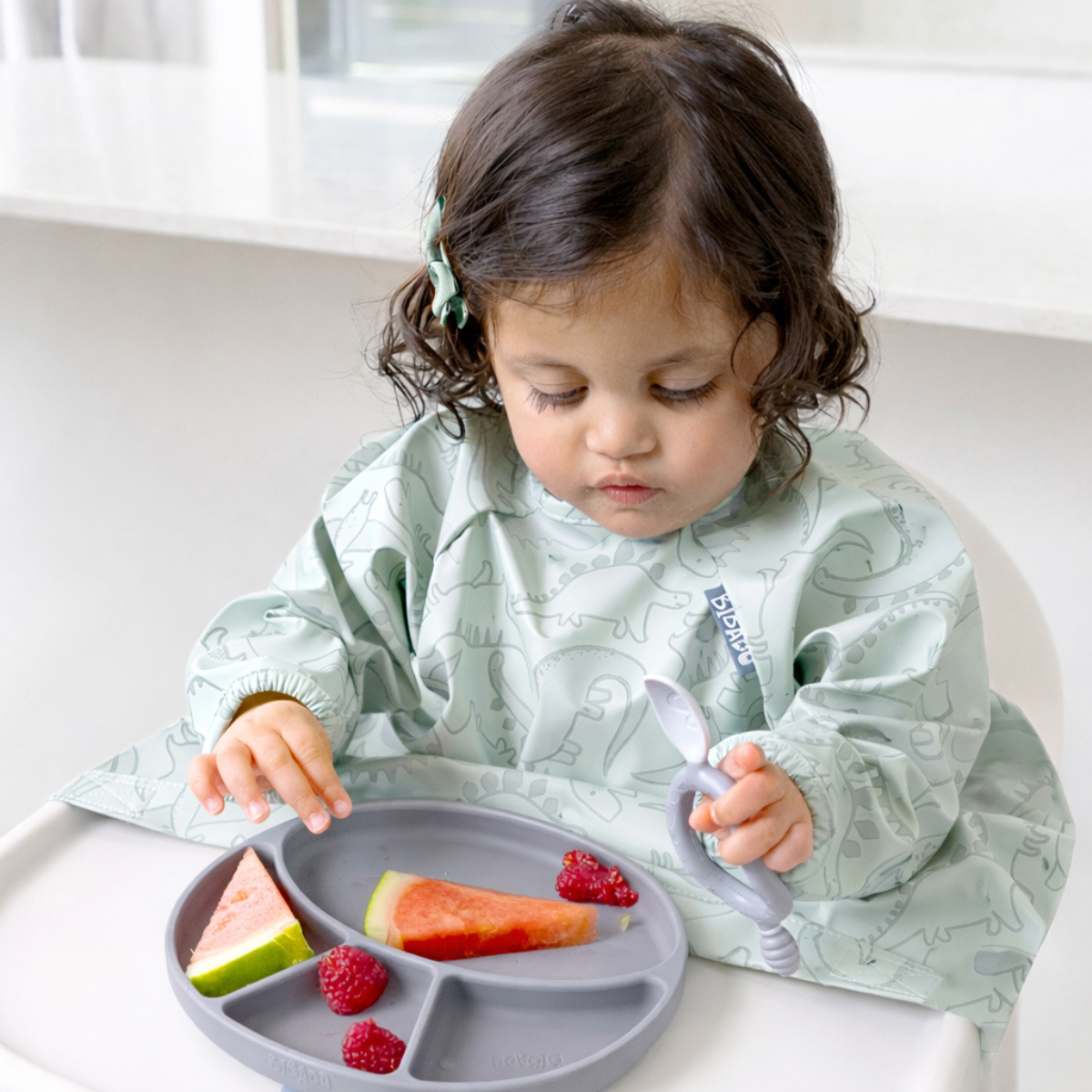 Child in a green Dino Coverall bib eating from a mist silicone divider plate with fruits. - Product image