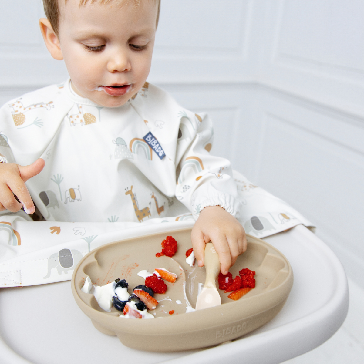 A baby wearing a white bib with a safari animal print sits in a high chair, reaching for food on a beige silicone plate. - Product image