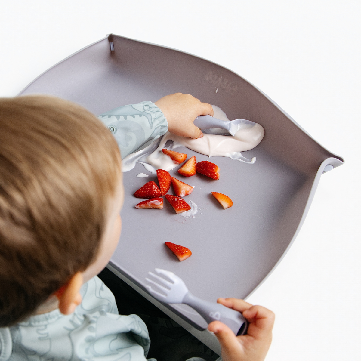 A child is shown from behind sitting at a blue-grey feeding mat, holding a matching baby spoon and fork while scooping yogurt and strawberry pieces against the side walls. - Product image