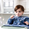 Baby in a blue space-themed bib holding a white Dippit™ multi-stage spoon with a textured dipper end.