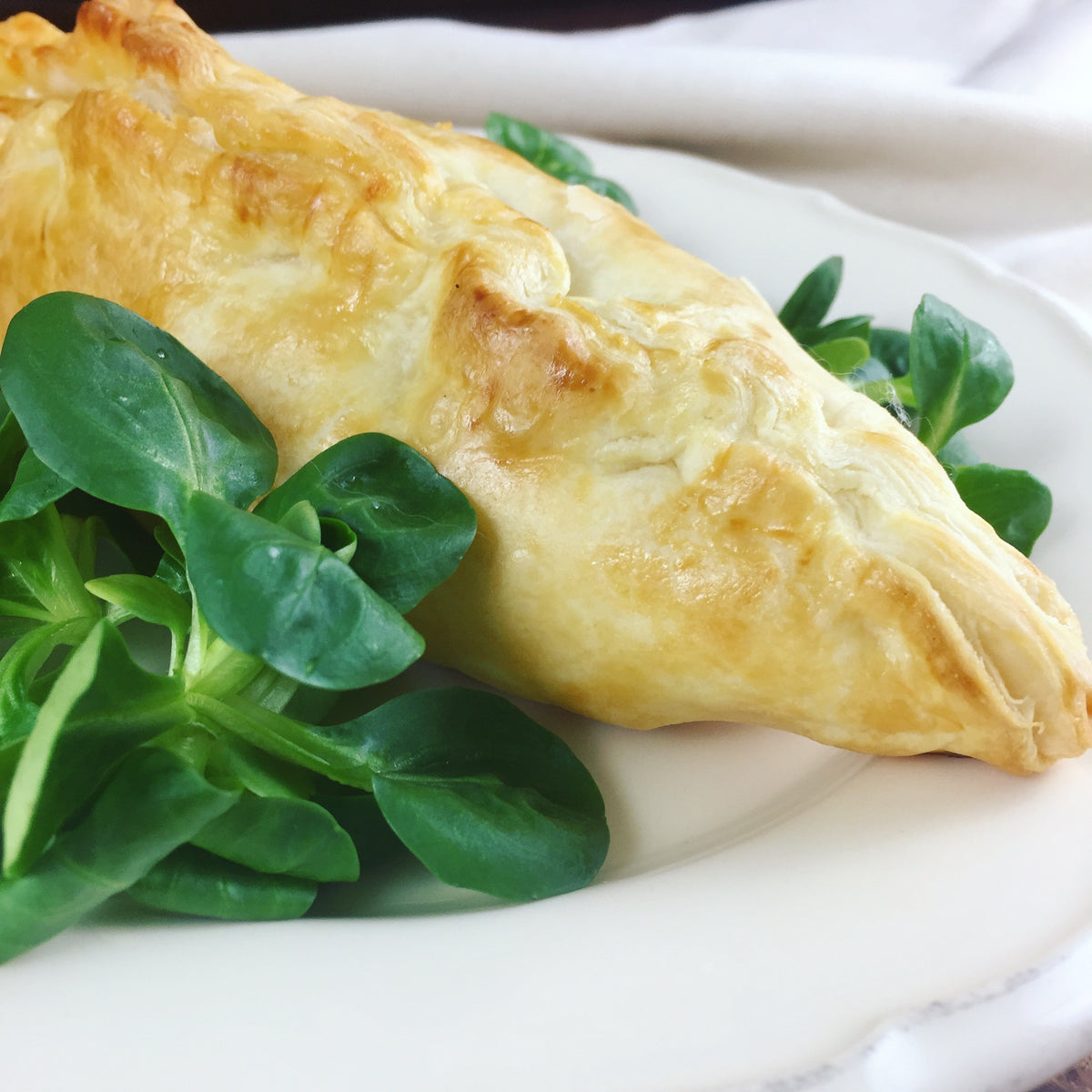 Golden-brown puff pastry turnover with a flaky crust, served on a white plate with fresh green lamb's lettuce. - Product image
