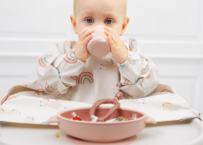 Baby in a rainbow-patterned bib drinks from a pink silicone cup, with a matching bowl of fruit. - image