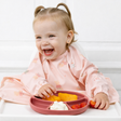 Child in a pink Coverall bib smiling whilst eating fruit from a blush silicone plate.