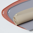 Close-up of a beige rolled silicone weaning mat with snap buttons resting on a blue-grey version fully rolled out, which is layered again over a pink mat on a clean white background.