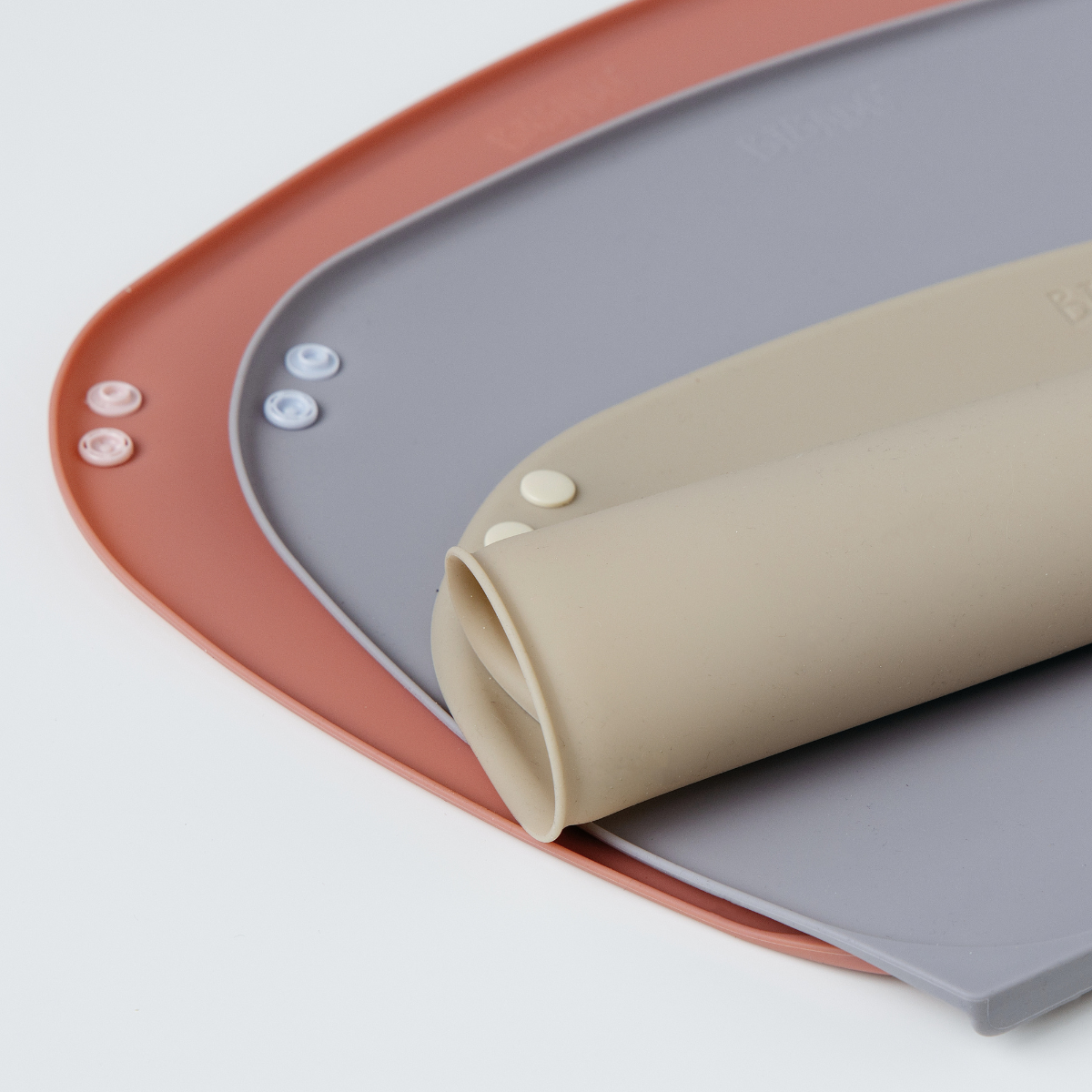 Close-up of a beige rolled silicone weaning mat with snap buttons resting on a blue-grey version fully rolled out, which is layered again over a pink mat on a clean white background. - Product image
