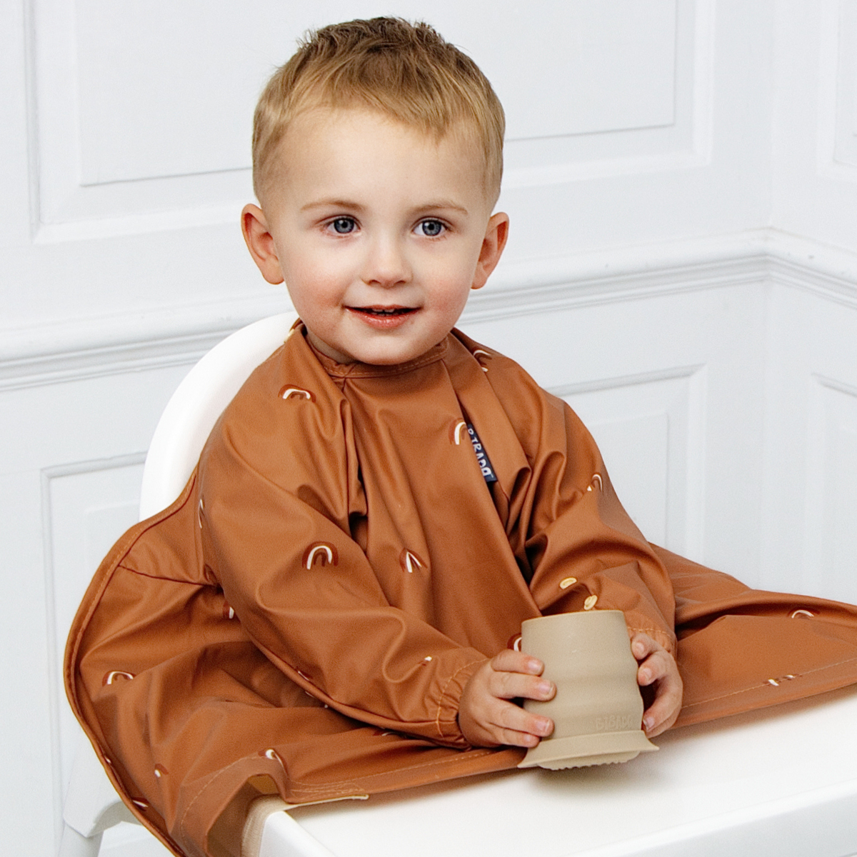 A young child sitting in a white high chair wearing a brown coverall bib with rainbow details, holding a silicone open cup. The background is a clean white panelled wall. - Product image