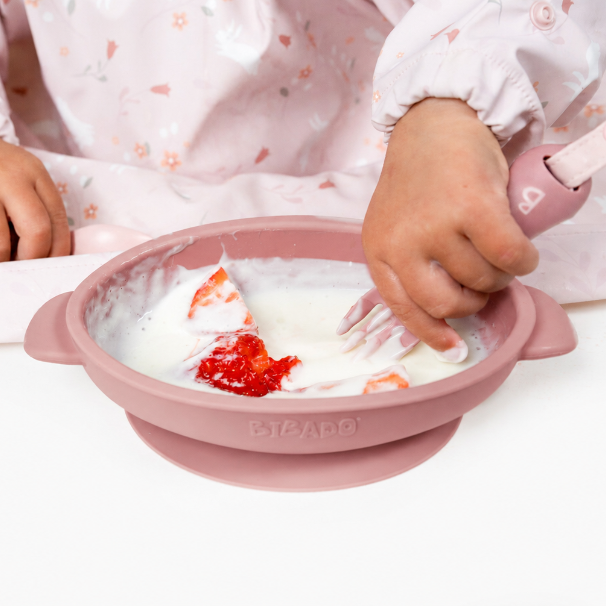 Baby in a pink floral bib using a pink silicone suction bowl with yogurt and strawberries, holding a pink spoon. - Product image