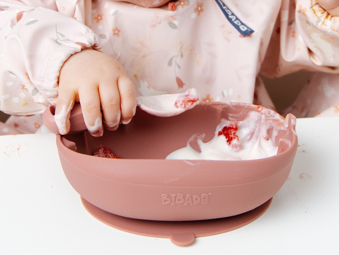 A baby wearing a light pink floral bib uses a dusty rose silicone spoon to scoop yogurt from a matching suction bowl. The brand name 'BIBADO' is visible on the bib and the bowl. - image