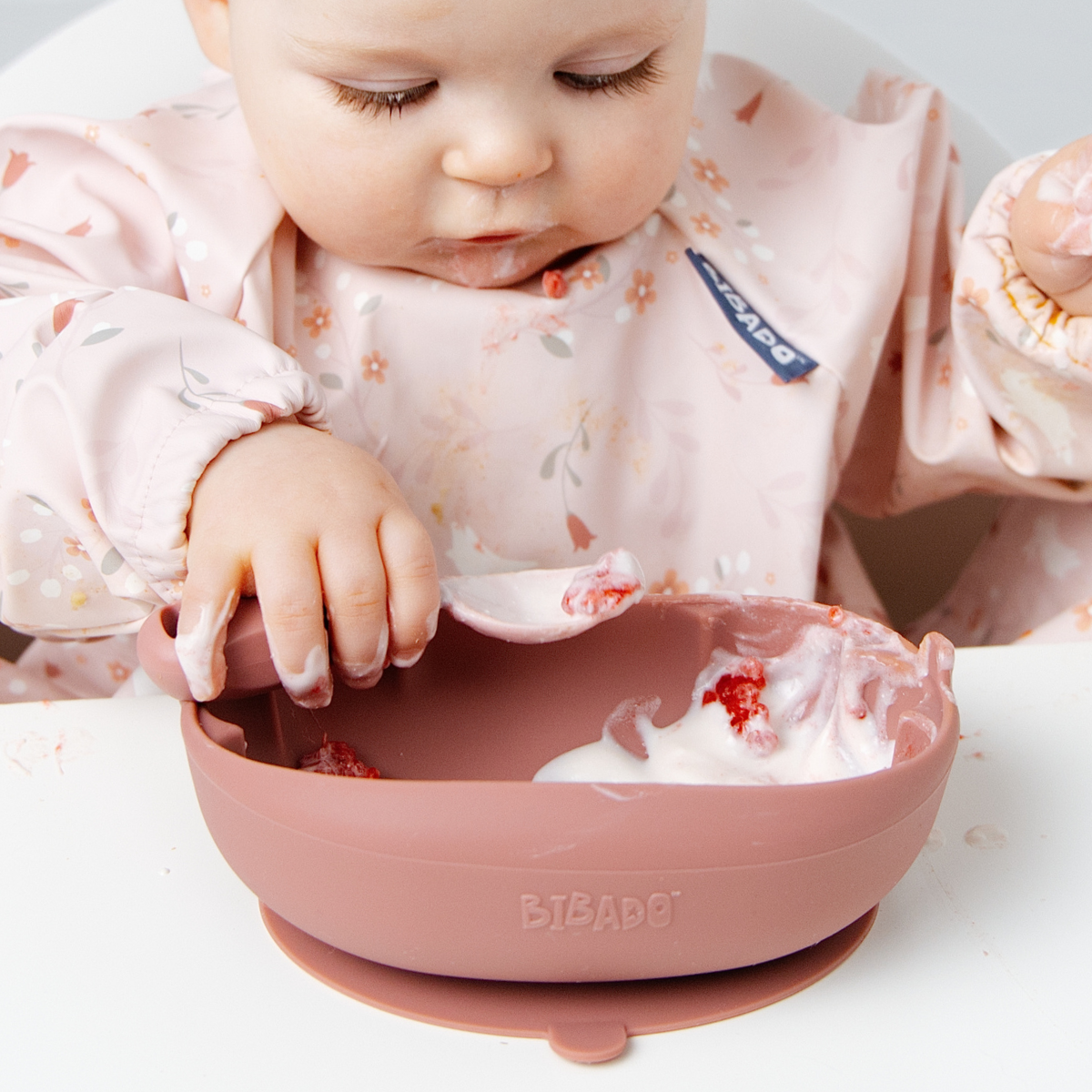 A baby wearing a light pink floral bib uses a dusty rose silicone spoon to scoop yogurt from a matching dusty rose suction bowl. The brand name 'BIBADO' is visible on the bib and the bowl. - Product image