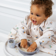 Happy baby in a high chair, wearing a patterned bib, playing with food in a light blue silicone suction bowl.